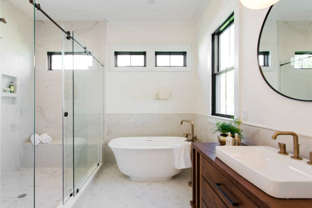 A modern bathroom featuring a sleek freestanding tub, glass-enclosed shower, wooden vanity with round mirror, brass fixtures, and bright natural lighting.